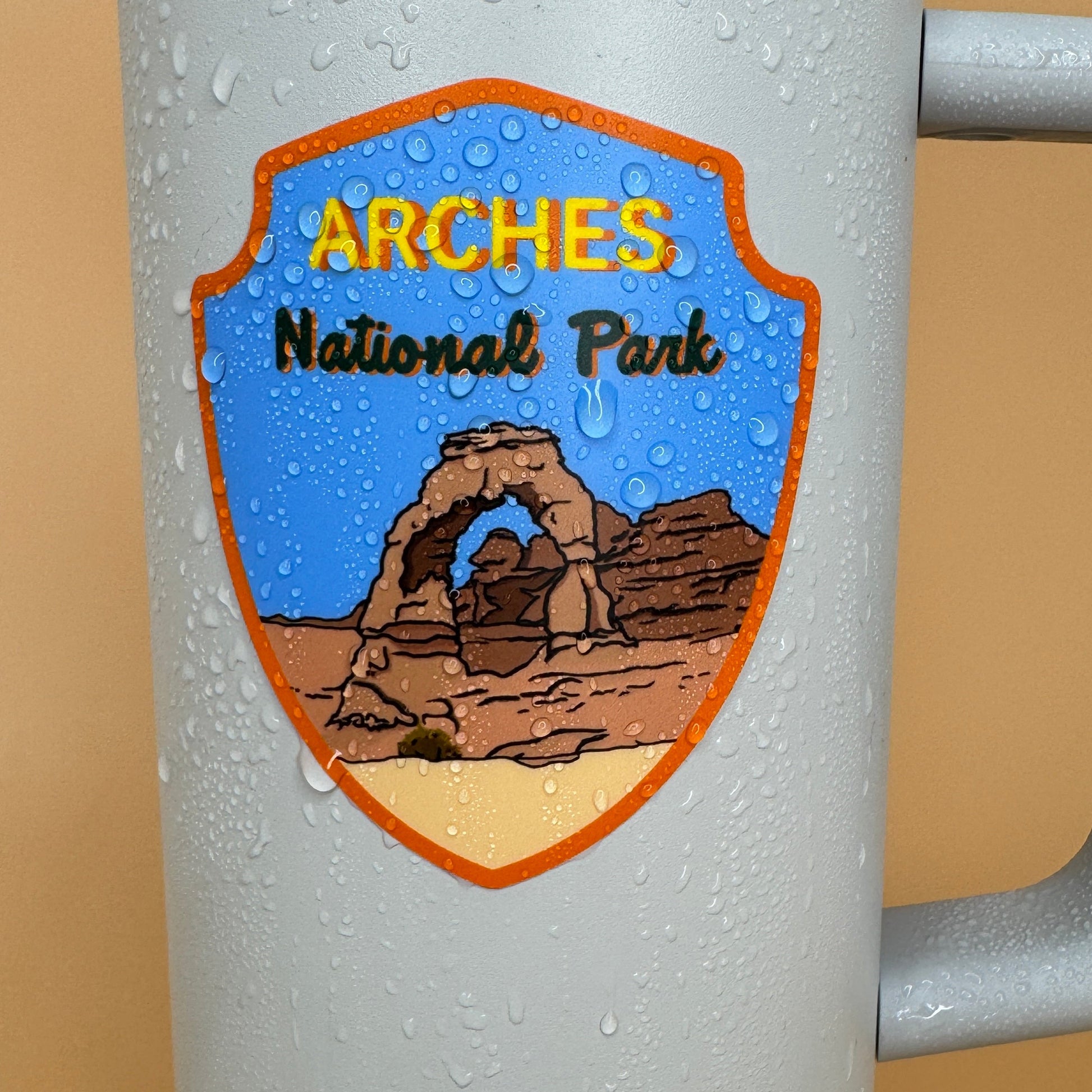 Grey tumbler with Arches National Park sticker covered in water droplets on a beige background